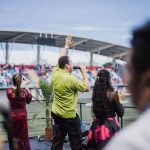 Grupo de artistas se apresenta em um estádio ao ar livre diante do público.
