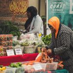 Duas mulheres organizam frutas e hortaliças frescas em uma banca de feira ao ar livre.