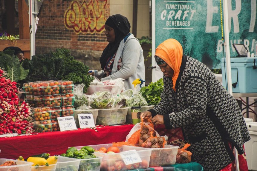Duas mulheres organizam frutas e hortaliças frescas em uma banca de feira ao ar livre.