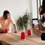 Grupo de jovens participa de um jogo criativo com copos vermelhos enquanto registra a atividade com um celular em ambiente interno.