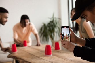 Grupo de jovens participa de um jogo criativo com copos vermelhos enquanto registra a atividade com um celular em ambiente interno.