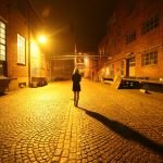 Mulher caminha sozinha por uma viela iluminada por postes durante a noite.