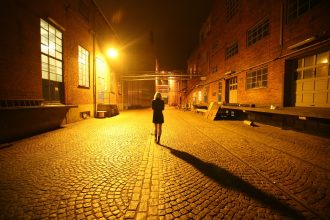 Mulher caminha sozinha por uma viela iluminada por postes durante a noite.